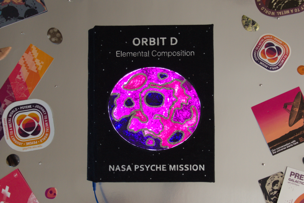 Psyche Mission Orbit D Notebook is the last in the series. The cover is a faux stained glass made of acrylic, and constructed with solder. The pattern represents elemental maps that assign an element a color and map it across the surface to see the element distribution. LED’s light up from behind giving a gemlike effect for the final touch.